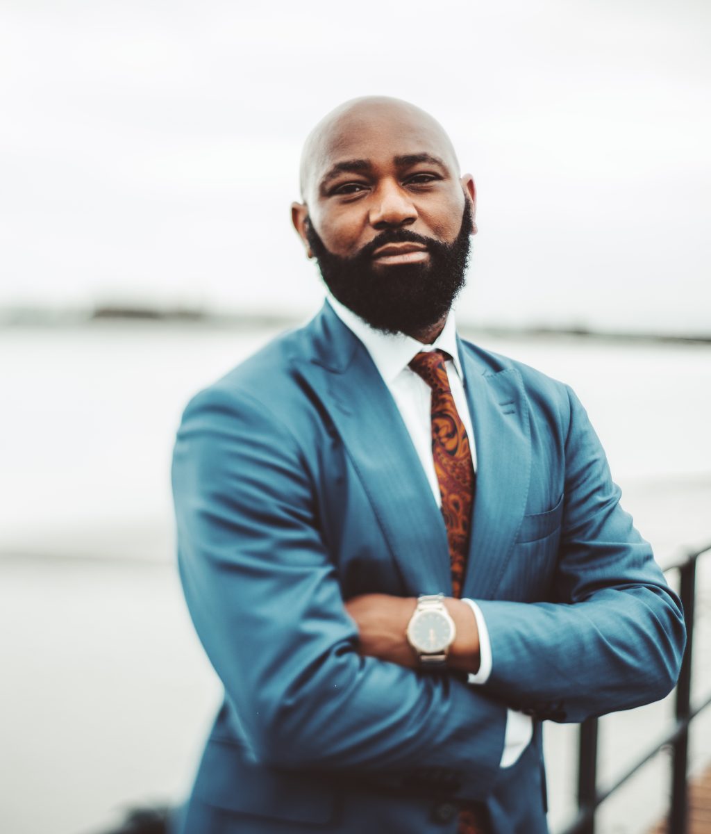 A true tilt-shift portrait of a successful adult African bald bearded man in a blue suit with tie and watch standing with hands crossed on an embankment near facing on an overcast day, selective focus
